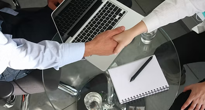 A professional handshake over a desk with a laptop and notebook, symbolising the successful placement of a qualified learning support assistant.