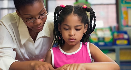 A learning support assistant with glasses gently helps a young girl in a pink shirt with her schoolwork, representing the ongoing professional development and support provided.