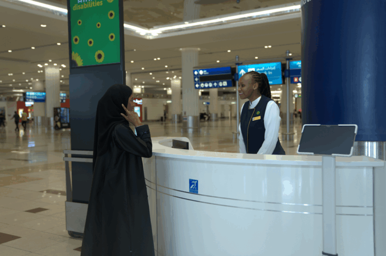 A female passenger, wearing a black abaya and hijab, stands at an airport information desk and speaks with a customer service assistant. The customer service assistant, a Black woman with her hair in a bun, smiles while listening to the passenger. The desk has a small blue sign with the international hearing loop symbol on it, indicating that it is fitted with the new technology for hearing-impaired travellers. This photo accompanies the news that Dubai Airports (DXB) has installed over 520 hearing loops across the airport to assist hearing-impaired travellers, marking the world's largest deployment of this technology.