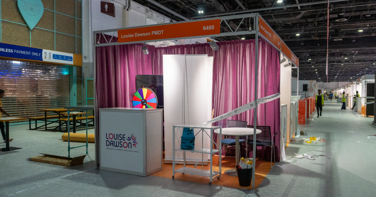 The Louise Dawson Inclusion stand at the AccessAbilities Expo in the Dubai World Trade Centre during setup, featuring purple curtains and branding, with a spin wheel and display areas ready for visitors.