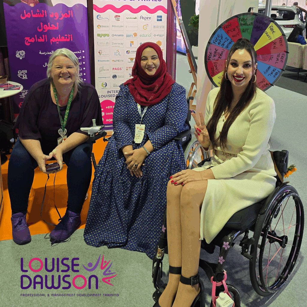 Louise Dawson and two other attendees in wheelchairs smiling for a photo at the Louise Dawson Inclusion stand, with a branded spin wheel in the background, showcasing positive connections made at the AccessAbilities Expo.