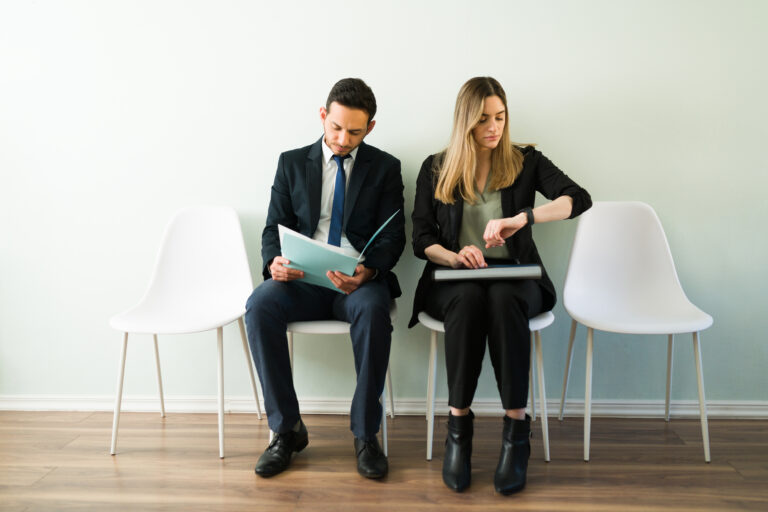 A picture of two job candidates waiting to be interviewed. One is checking her watch, the other is checking his notes.