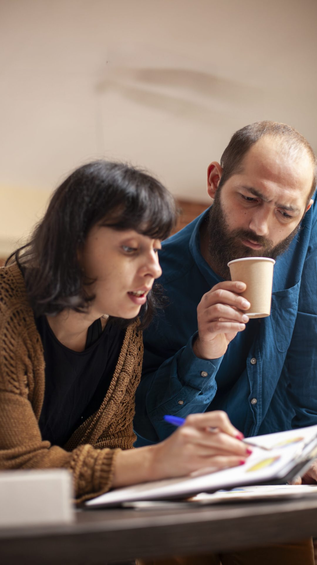 Bearded man in casual business clothing holds coffee cup and reviewing financial paperwork with female coworker. Male employee listens to businesswoman explaining company research data on clipboard.